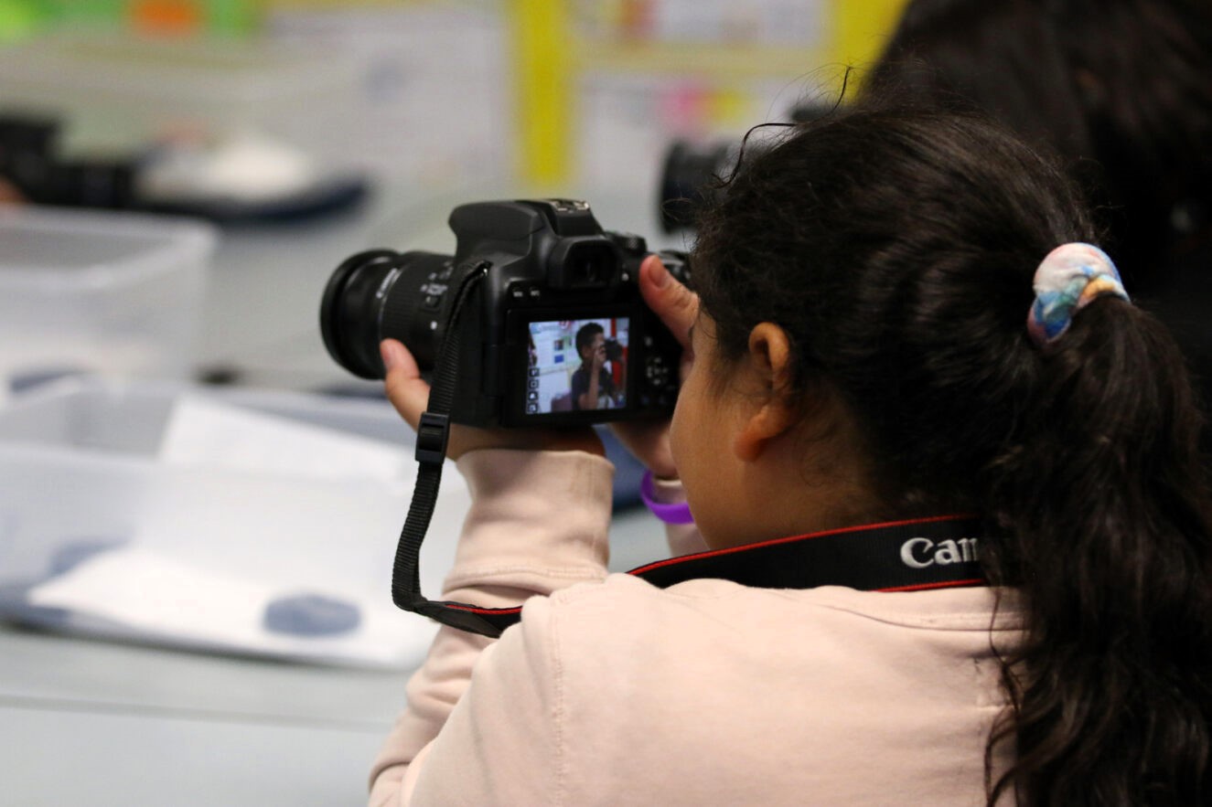 A girl takes a photo with another student in her class appearing on her camera screen.