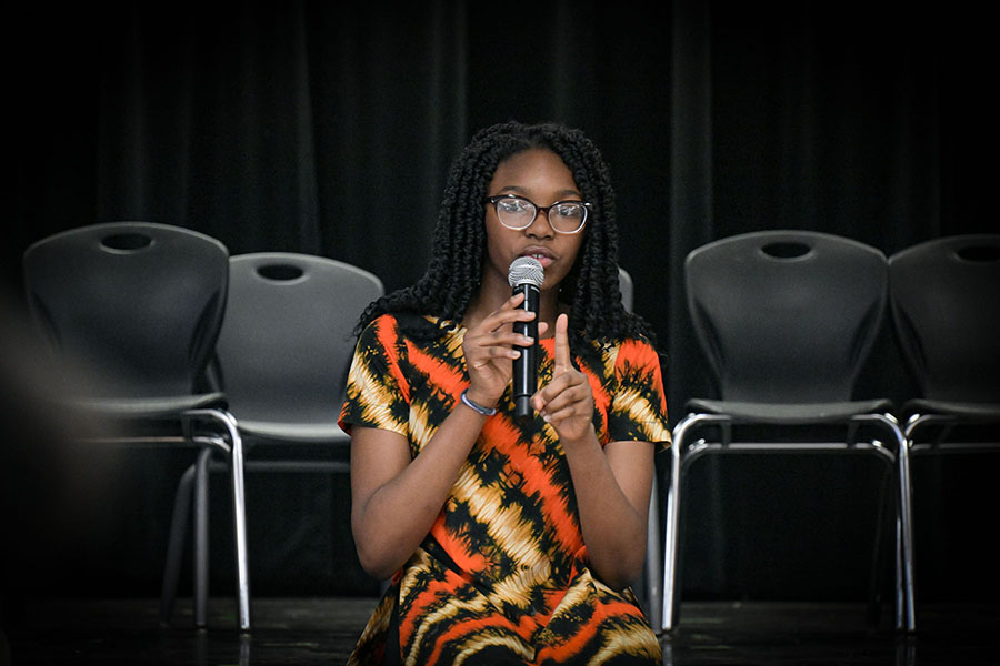 A teen girl sits on the stage talking to students
