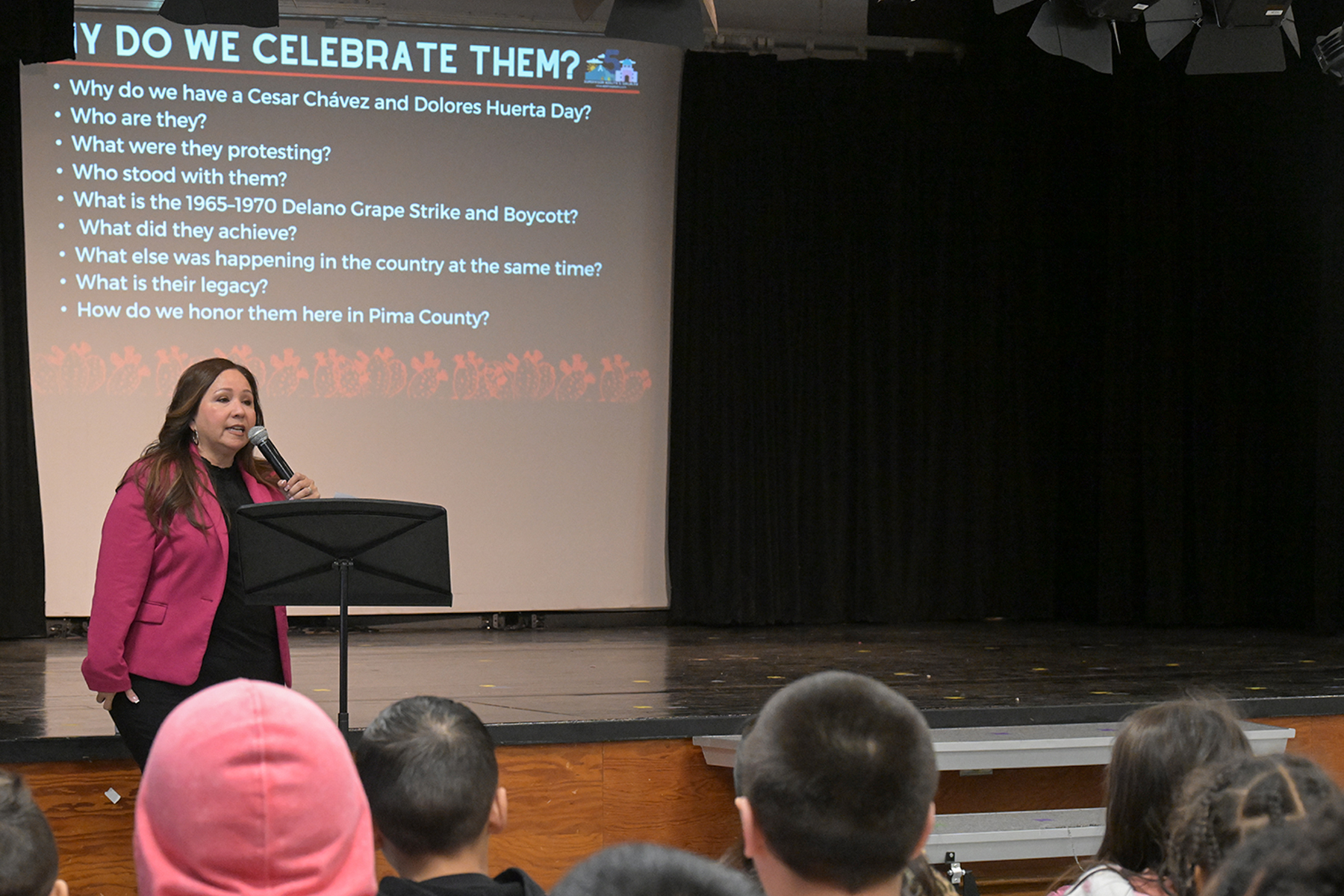 A woman in a pink jacket talks in a mic to students with a screen behind her with talking points about Chavez and Huerta