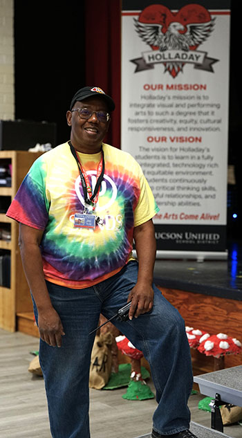 A man in a tie-dye shirt stands in front of a Holladay banner