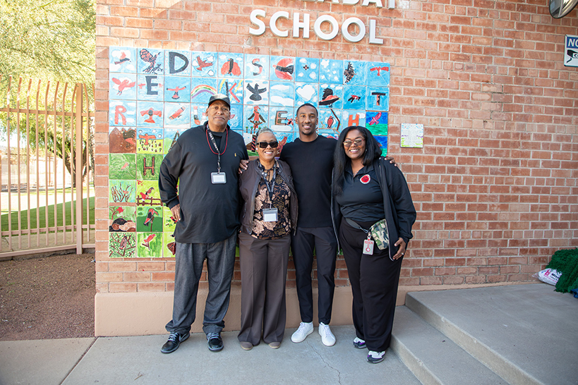 Two men and two women smile outside in front of the school's mural