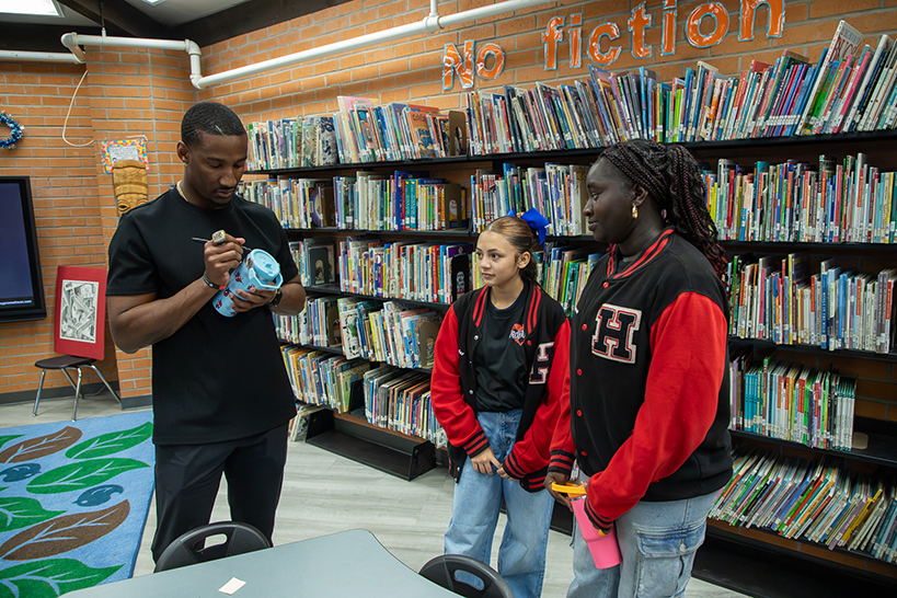 A man signs a blue water bottle as two girls in red and black letterman jackets look on