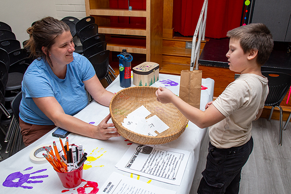 A woman in a blue shirt holds out a basket for a little boy to grab a piece of paper out of