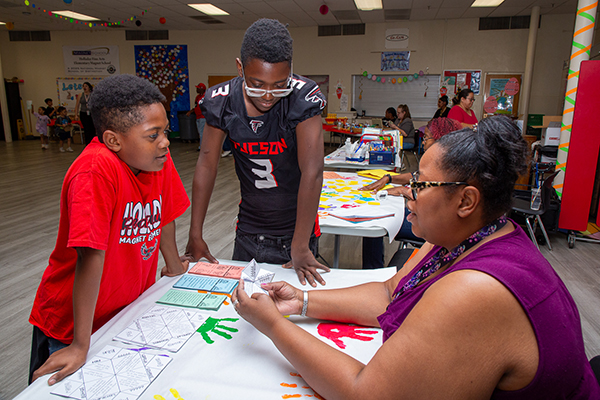 A woman holds up a paper fortune teller as she talks to two boys