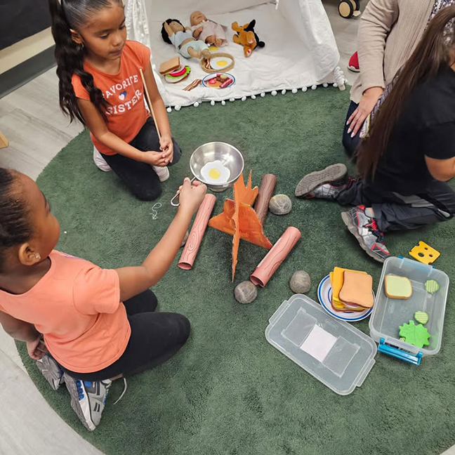 Two little girls pretend to cook an egg over the campfire, with a tent in the background